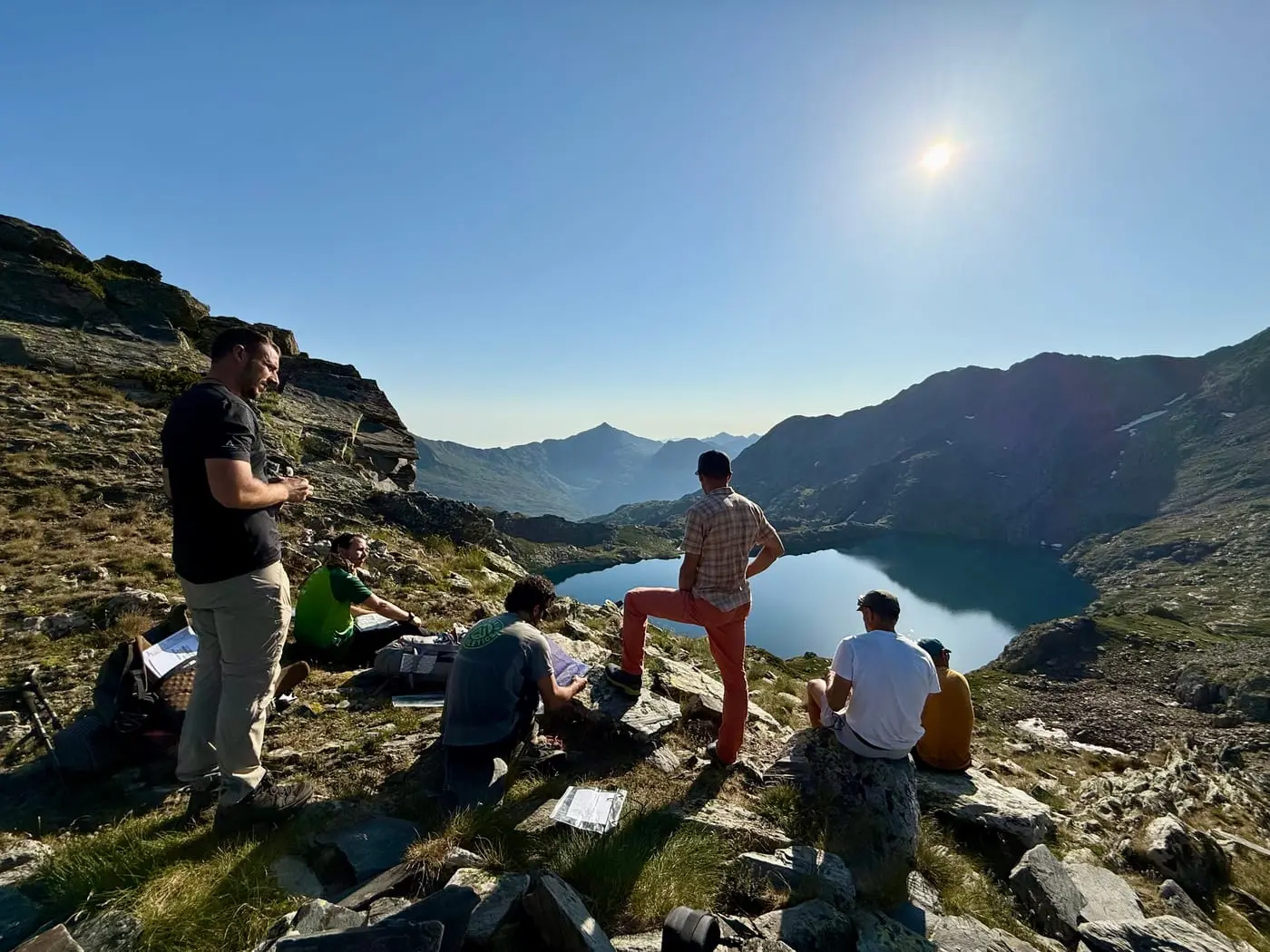 A group resting on a grassy shoulder high above a blue mountain lake, with Pyrenean ridges running off into the sun.